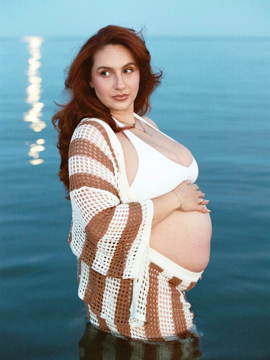 A pregnant woman with long red hair stands in the ocean wearing a white bikini top and a brown and white striped crochet cover-up.