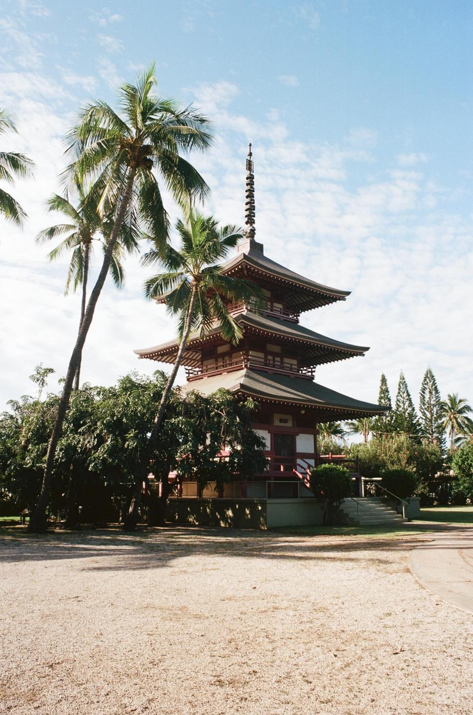 A red and white pagoda with multiple tiered roofs stands among palm trees and other foliage under a blue sky with scattered clouds.