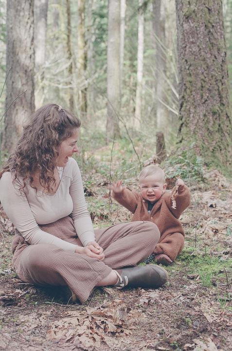 A woman and a baby sit on the ground in a forest.