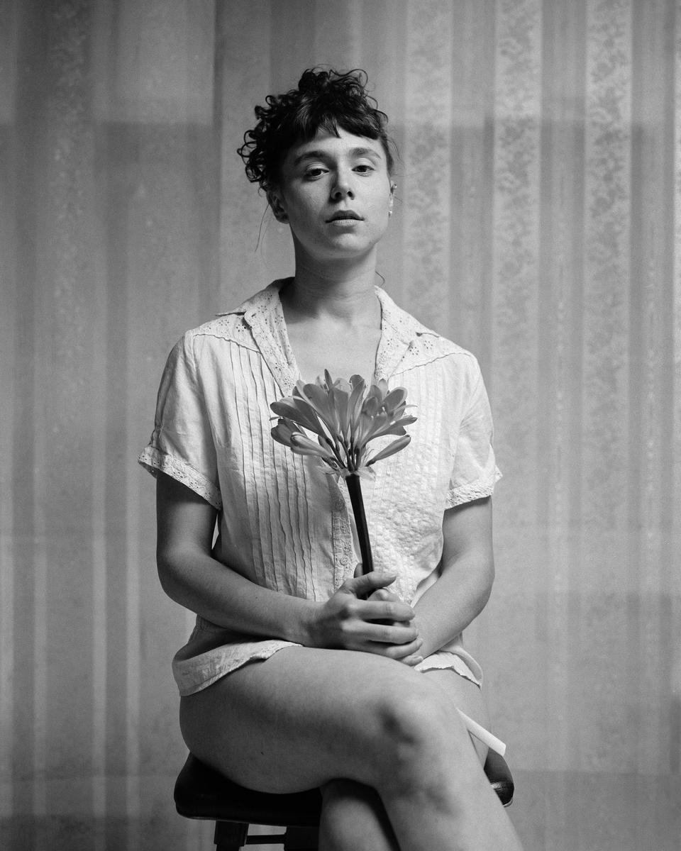 A woman with curly hair sits on a stool holding a flower in front of a patterned curtain.
