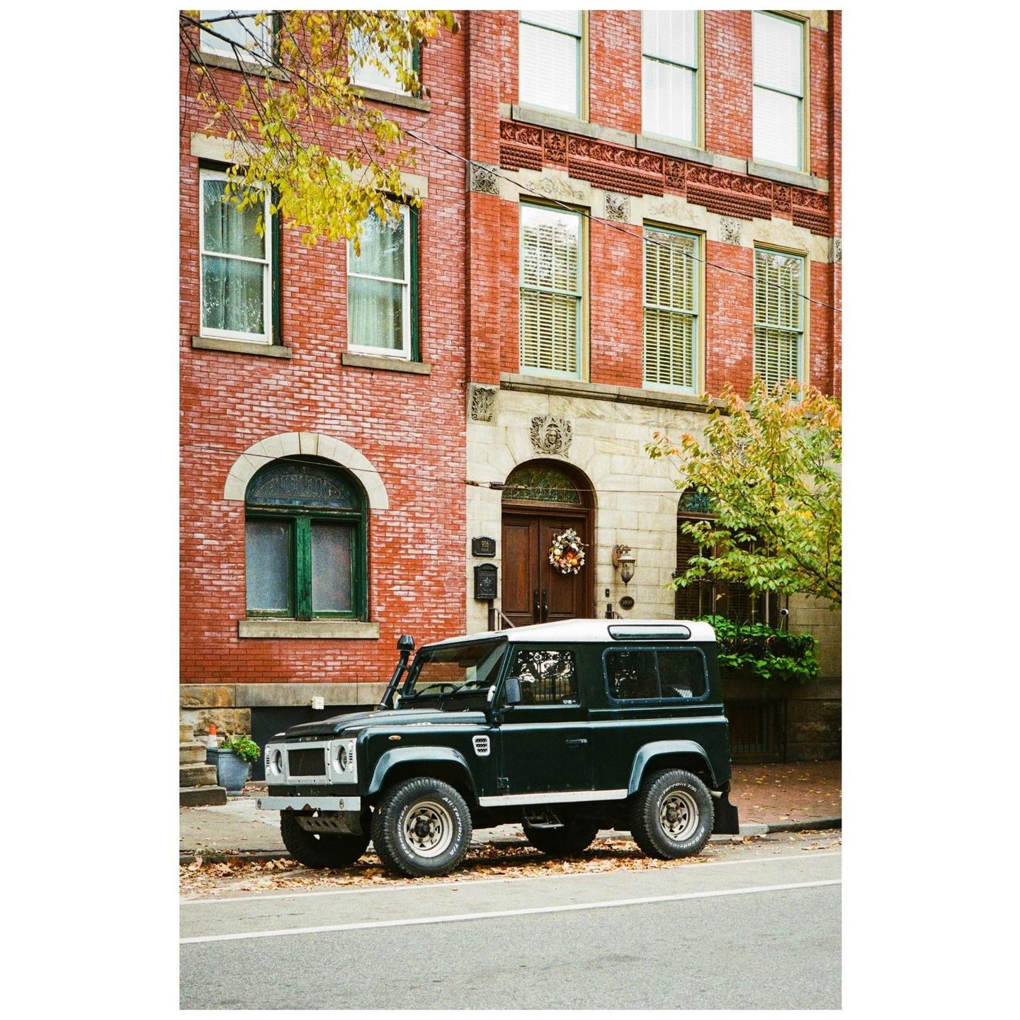 A dark green sport utility vehicle is parked on a street in front of a brick building with a wreath on the door.