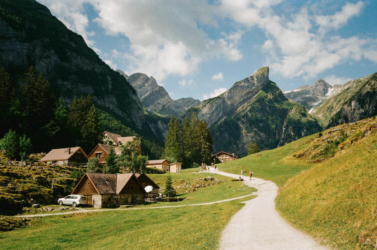 A winding path leads through a green valley with houses nestled among trees, surrounded by towering mountains under a cloudy sky.