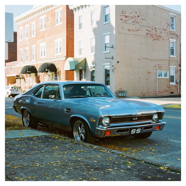 A blue vintage car is parked on a street with fallen autumn leaves in front of brick buildings.