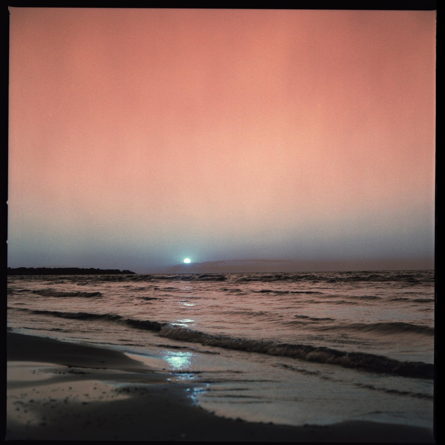 The sun sets over a body of water with a sandy shore in the foreground.