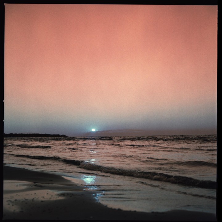 The sun sets over a body of water with a sandy shore in the foreground.