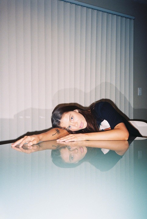 A woman with a palm tree tattoo on her arm is resting her head on a reflective table.