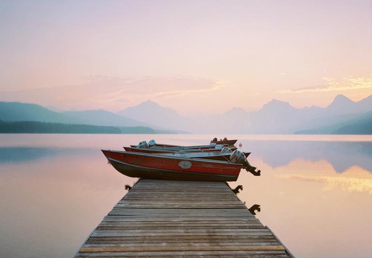 Several red boats are docked on a wooden pier extending into a calm lake with mountains in the background under a pastel sky.