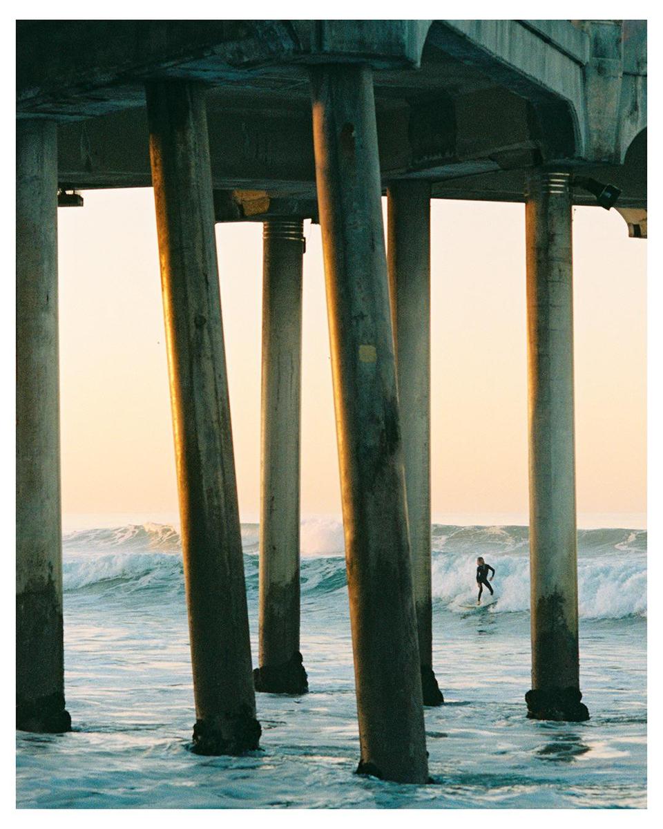 A surfer rides a wave between the pillars of a pier at golden hour.