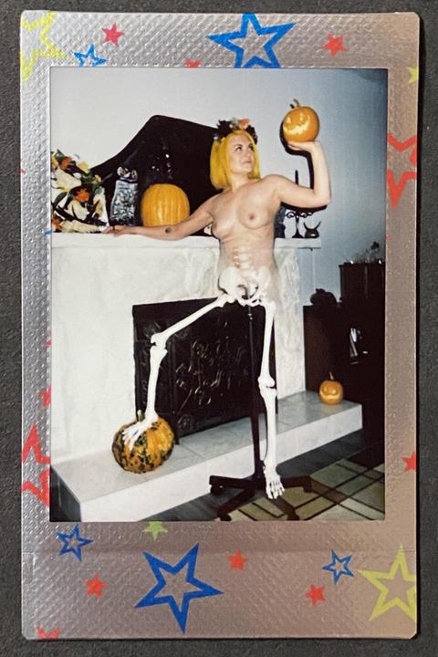 A woman with yellow hair and a skeleton costume holds a pumpkin while standing in front of a fireplace.