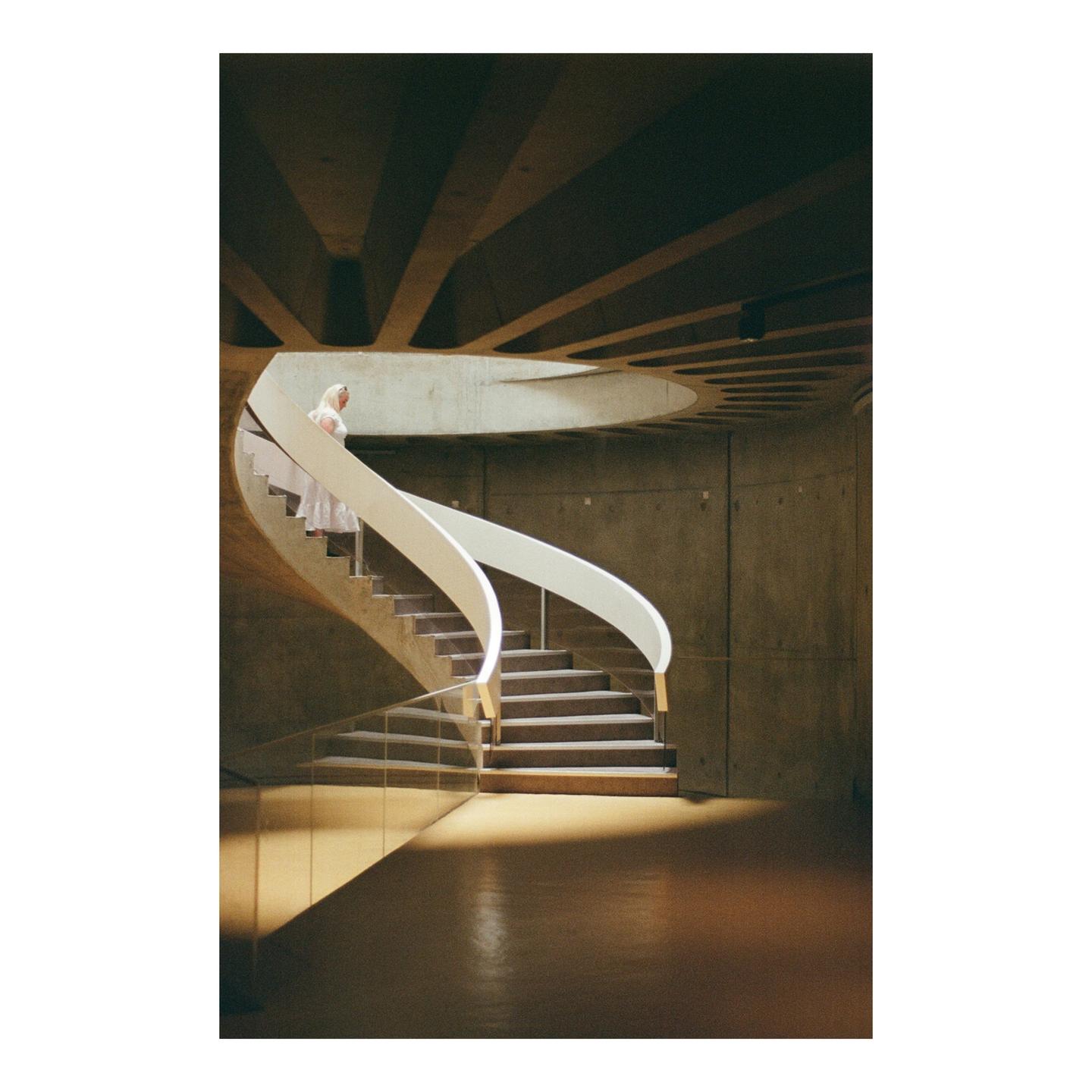 A woman in a white dress descends a spiral staircase with a white railing in a building with concrete walls and ceiling.
