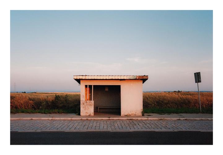 A small, white bus stop shelter with a bench inside stands beside a paved road and a field of tall, dry grass under a clear sky.
