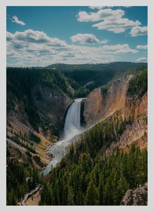 A large waterfall cascades into a river in a deep canyon surrounded by pine trees under a blue sky with clouds.