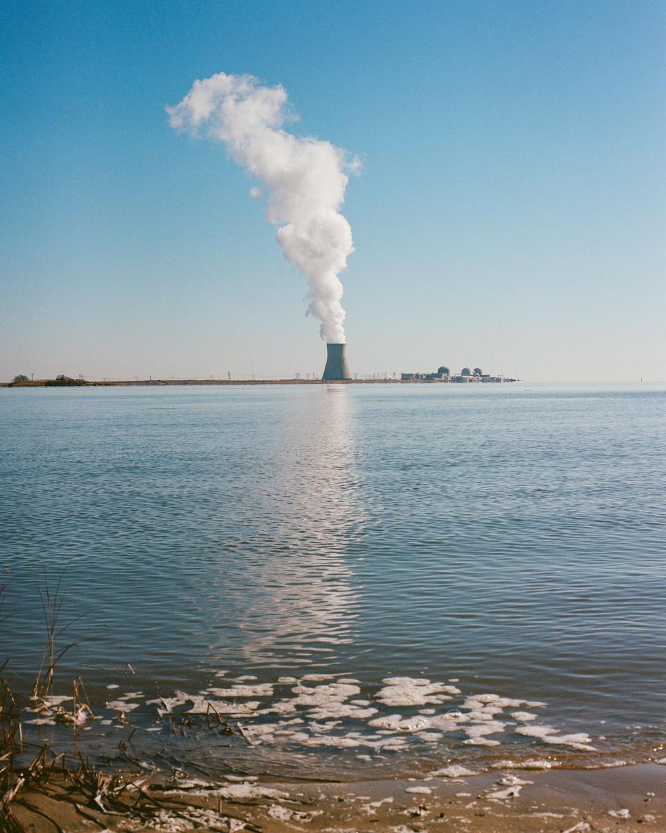 A power plant with a tall smoke stack emitting a large plume of smoke stands across a body of water under a clear sky.