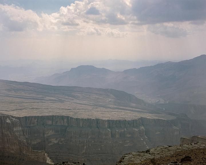 A vast canyon with a flat plateau on one side and layered mountains in the distance is visible under a cloudy sky.