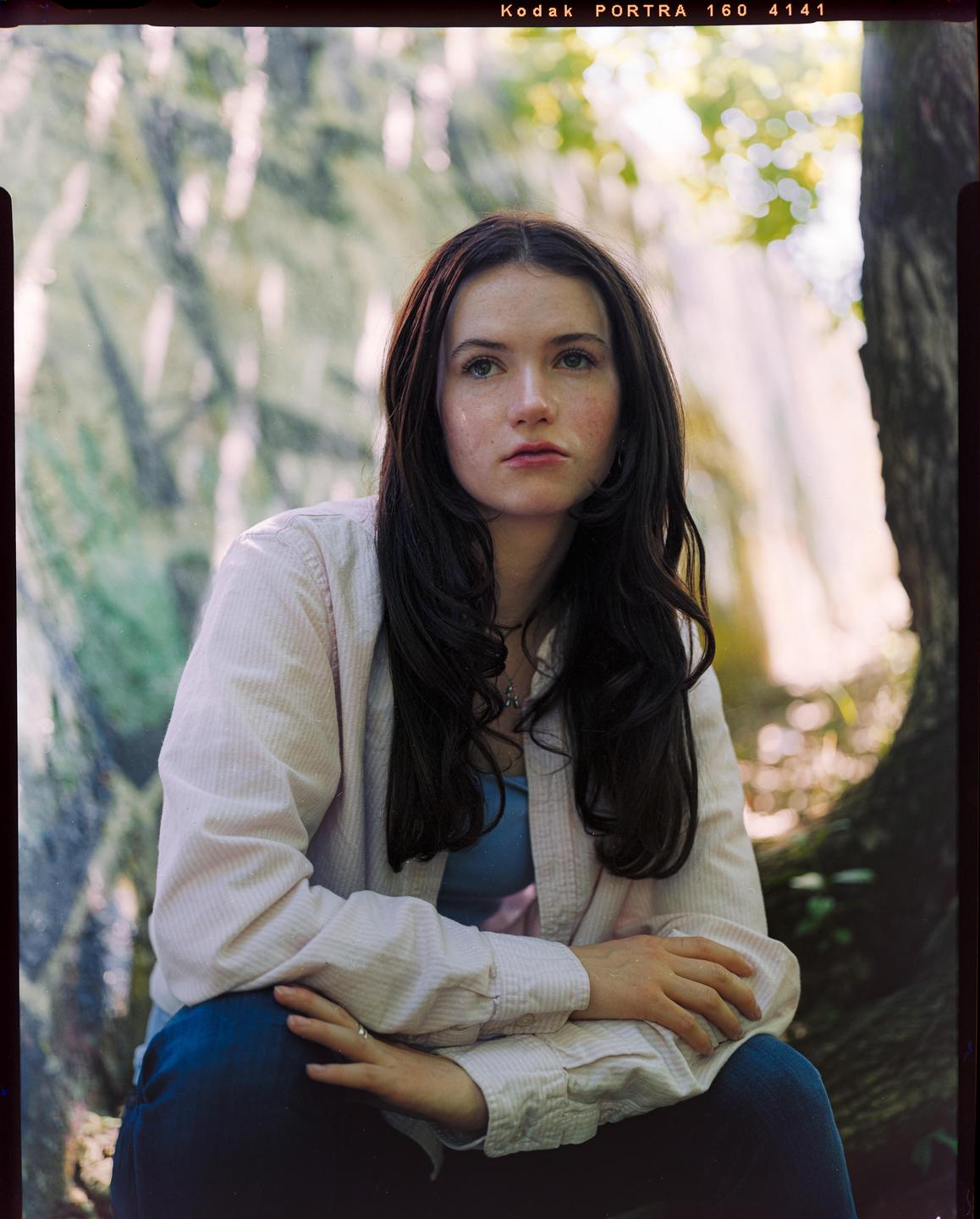 A woman with long dark hair sits outdoors, looking upwards with a thoughtful expression.