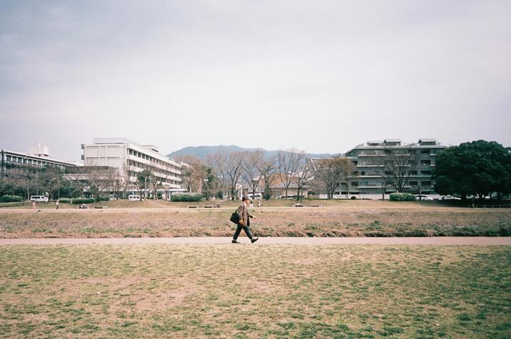 A man walks across a grassy field with buildings and trees in the background.