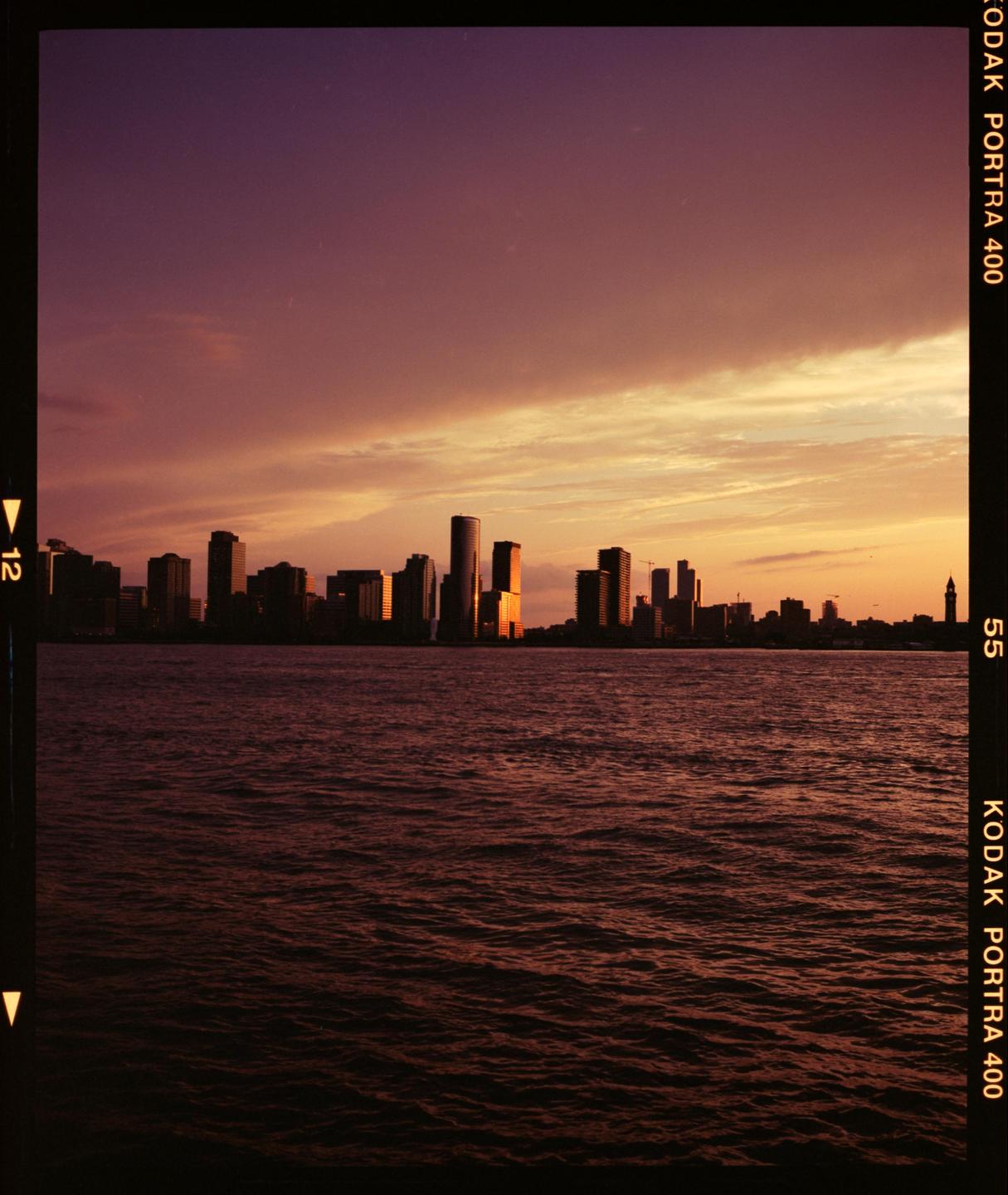 A city skyline is silhouetted against a colorful sunset sky over a body of water.
