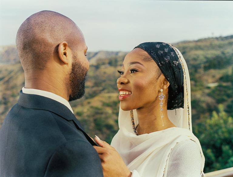A woman in a headscarf smiles at a man in a suit, with hills in the background.
