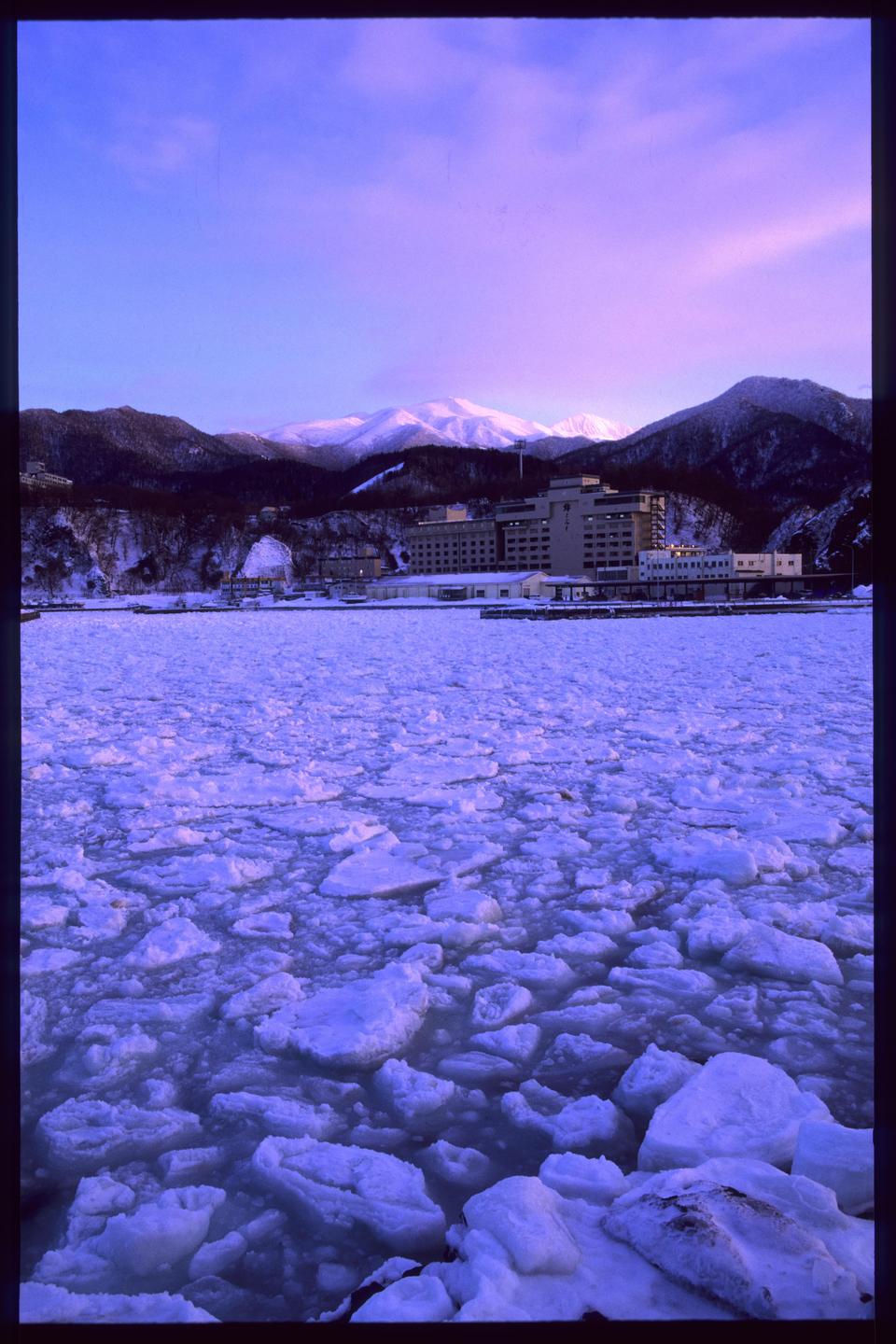 A body of water filled with drift ice is in the foreground, with a large building and snow-capped mountains in the background under a purple sky.