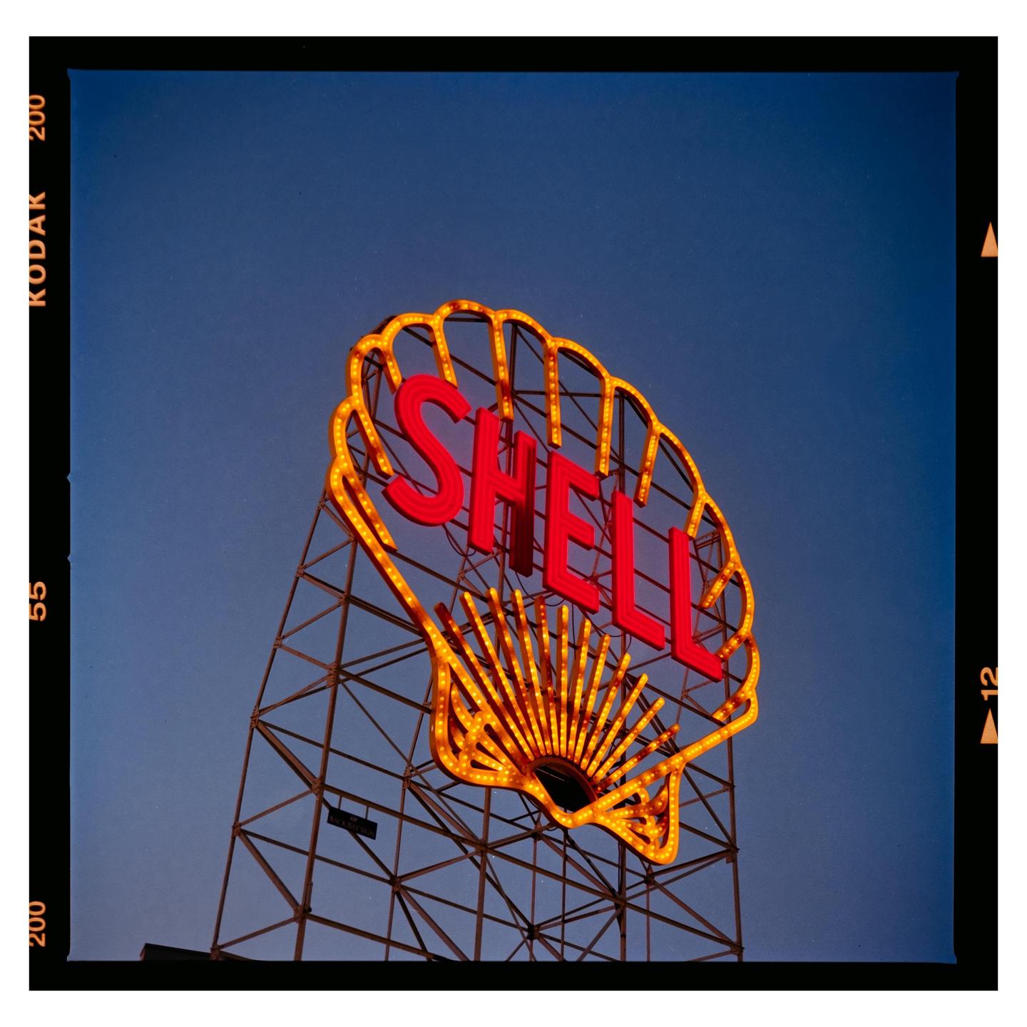 A large Shell sign with red and yellow neon lights stands against a clear blue sky.