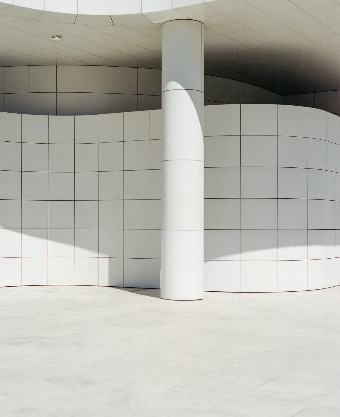 A white column stands in front of a curved tiled wall, casting a shadow on the ground.