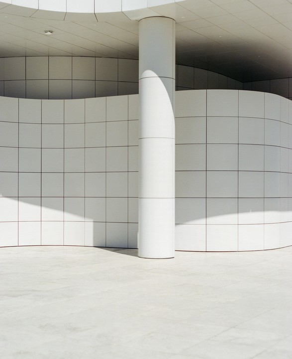 A white column stands in front of a curved tiled wall, casting a shadow on the ground.