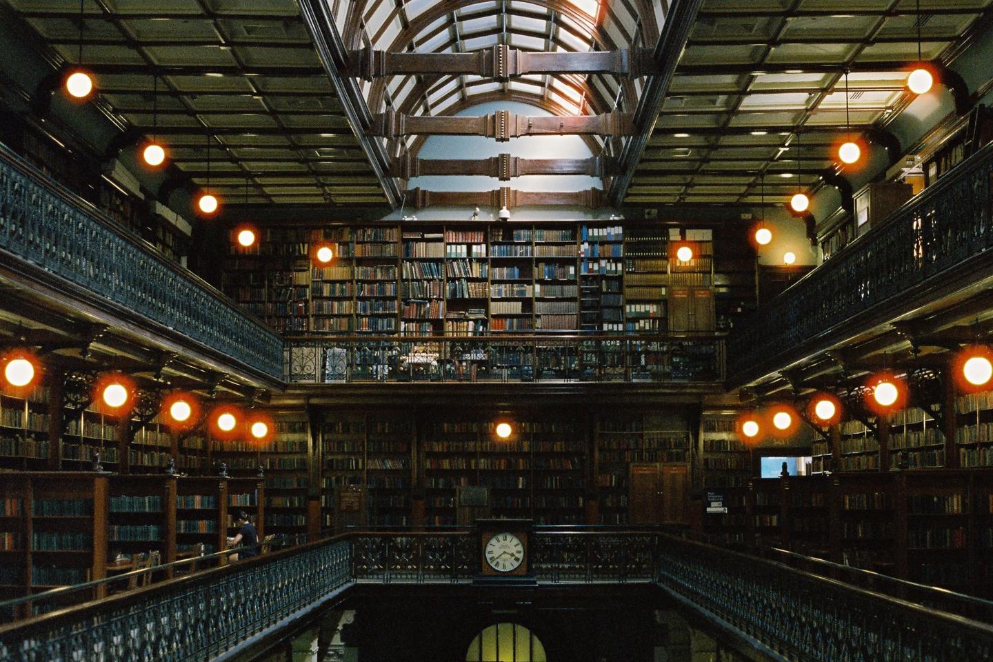 A grand library interior features multiple levels of bookshelves, ornate railings, and glowing spherical lights, with a large clock visible on the lower level.
