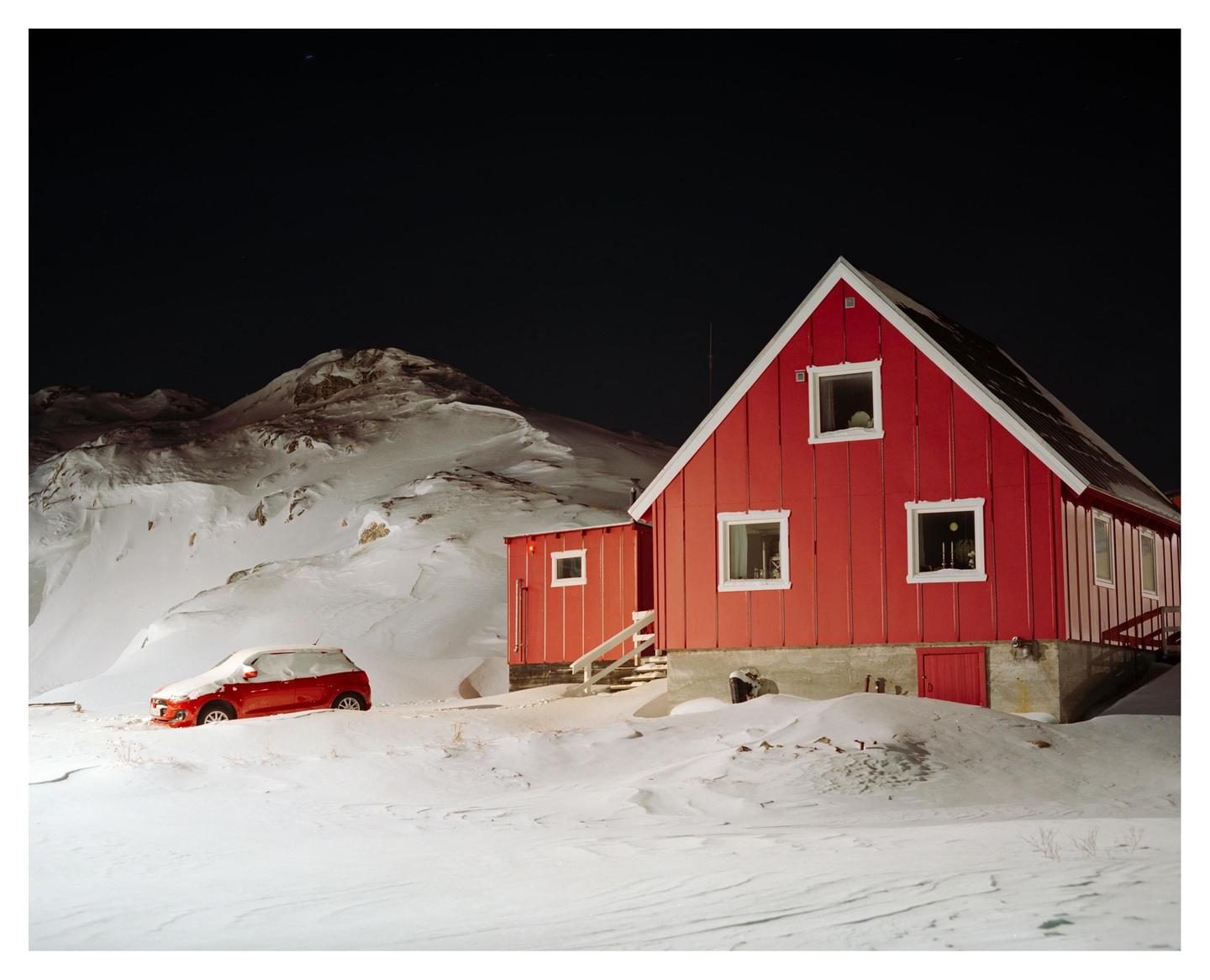 A red house and a red car are surrounded by snow at night with mountains in the background.