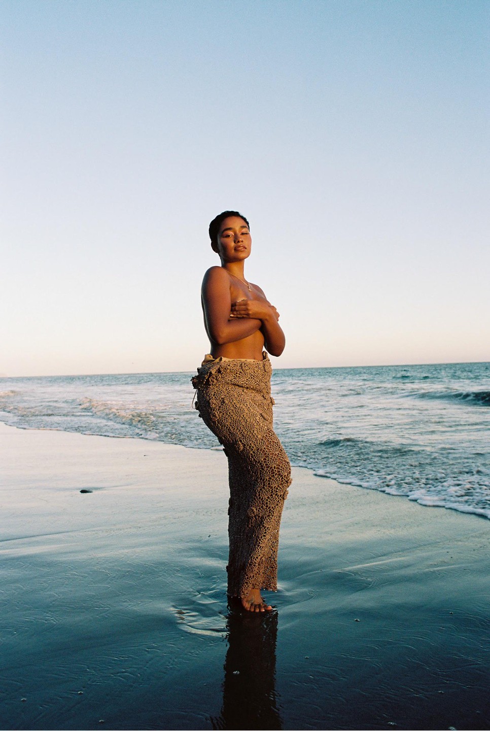 A woman stands in the shallow water of a beach, covering her chest with her arms.