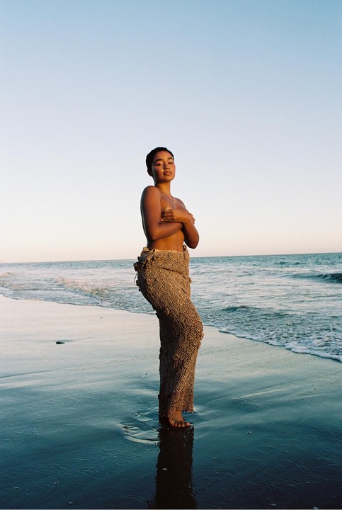A woman stands in the shallow water of a beach, covering her chest with her arms.