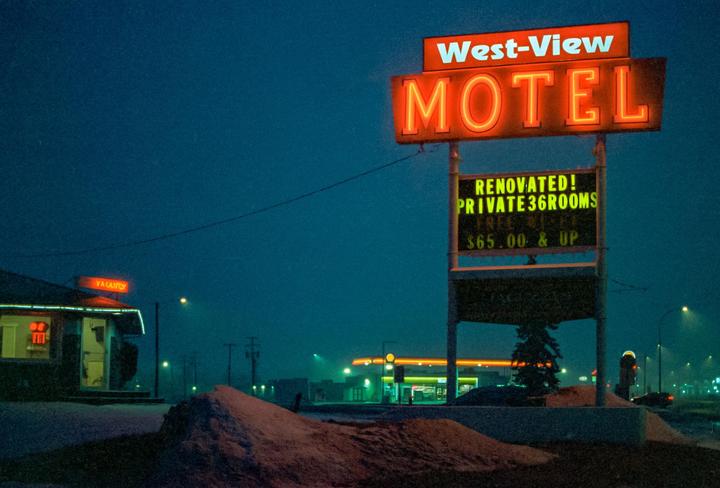 A neon motel sign stands in front of a building with a "vacancy" sign, with snow on the ground and a gas station in the background.
