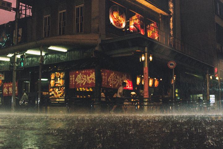 Rain falls on a street in front of a brightly lit building with red banners and lanterns at night.