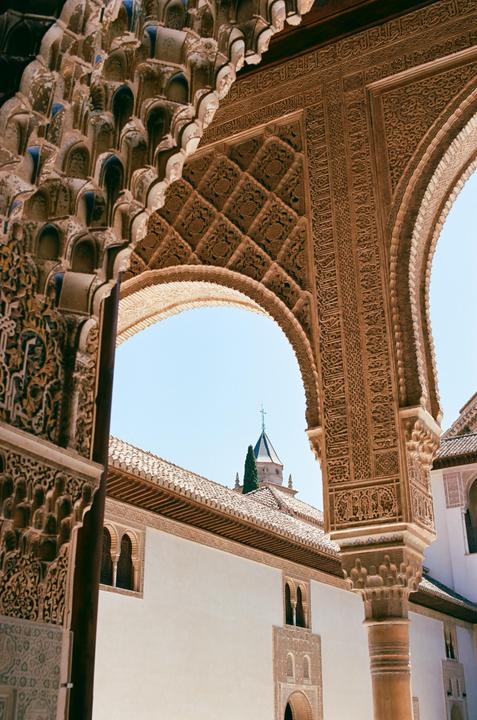 An ornate archway frames a view of a building with a tiled roof and a distant tower against a clear sky.