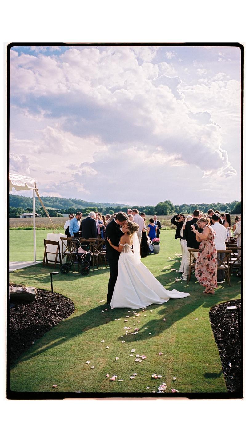 A bride and groom kiss on a grassy lawn surrounded by wedding guests under a cloudy sky.