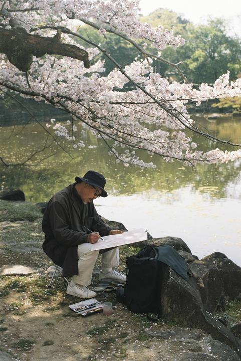 A man wearing a hat and glasses sits on a stool by a body of water, painting on a canvas under a blossoming tree.