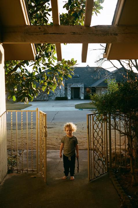 A child stands barefoot on a porch holding a stick, looking out through an open gate towards a house across the street.