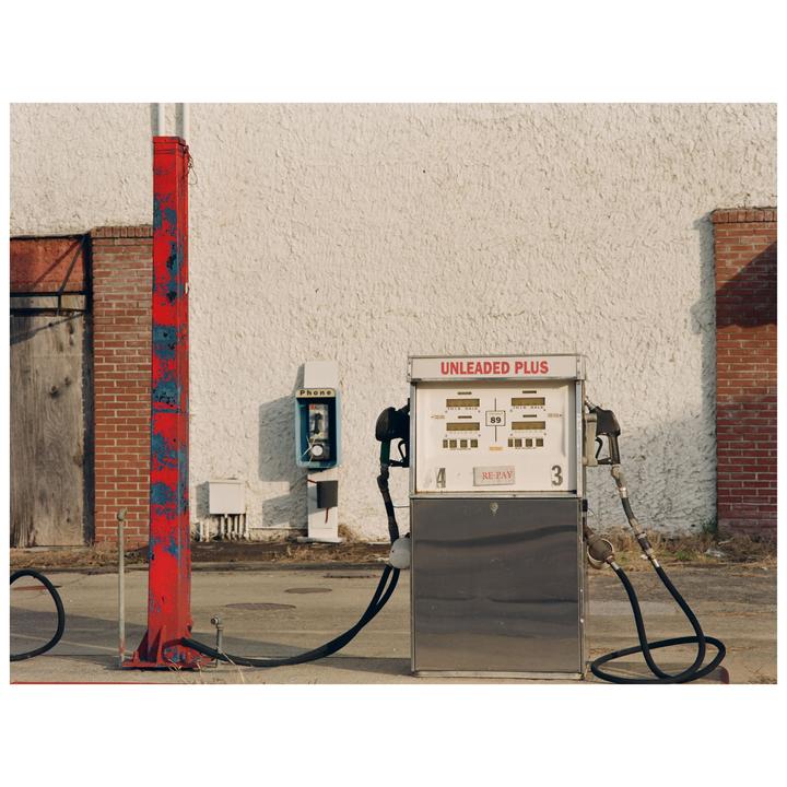 A gas pump and a payphone stand in front of a white building with a red pole in the foreground.