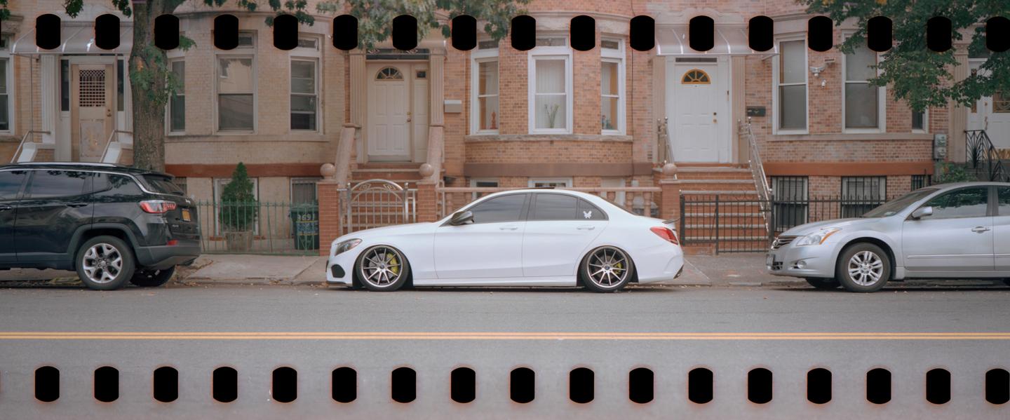 A white sedan with custom wheels is parked on a residential street in front of brick buildings.