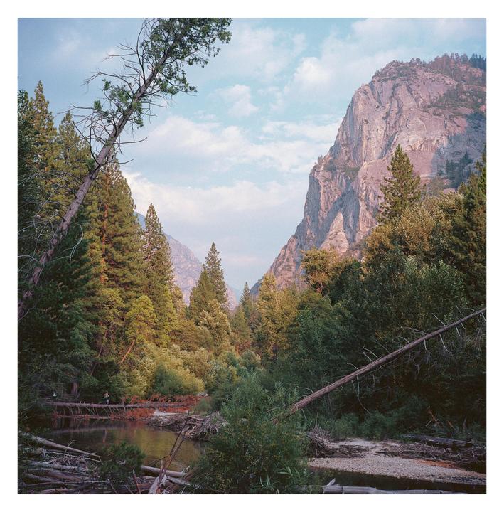 A river flows through a forest with mountains in the background under a cloudy sky.