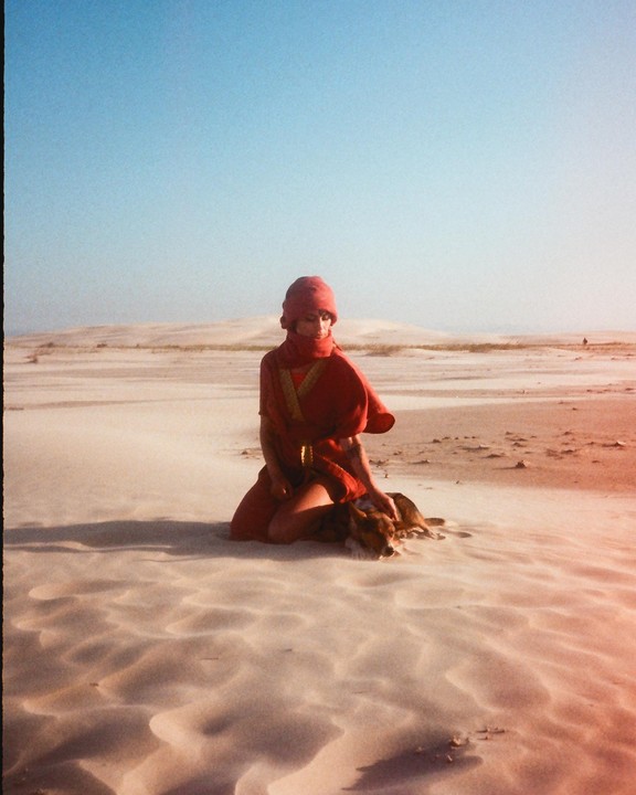 A woman in a red dress and head covering kneels in a desert landscape, petting a dog.