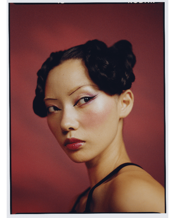 A woman with elaborate hair and makeup looks over her shoulder against a red background.