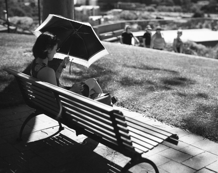 A woman sits on a park bench holding an open umbrella over her head, with other people in the background.
