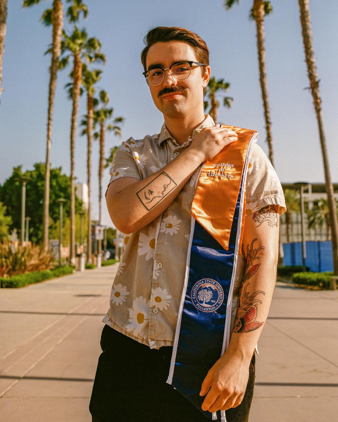 A man wearing a daisy-patterned shirt and a graduation stole stands on a paved path lined with palm trees.