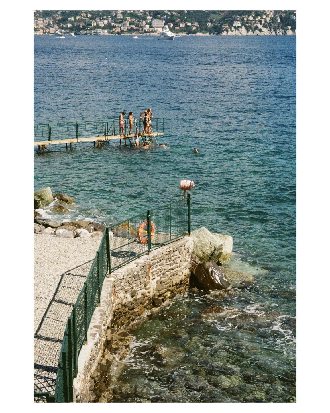 A group of people are swimming and standing on a pier in the sea with a town and hills in the background.