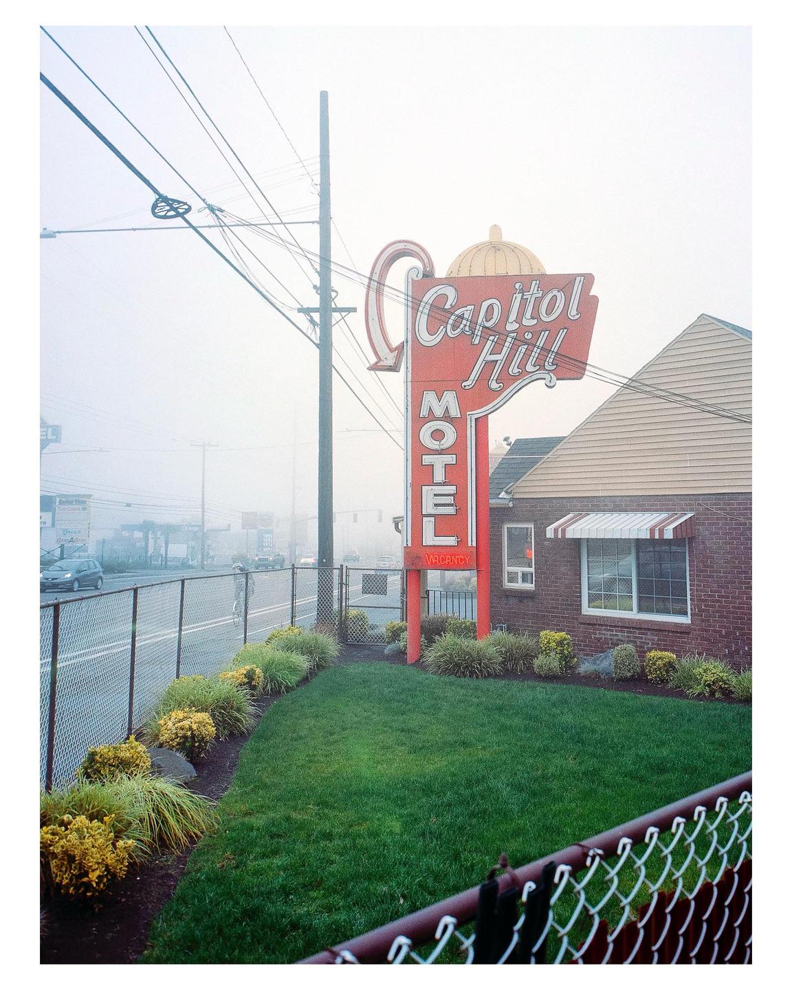 A red and white motel sign stands in front of a brick building with a green lawn and a chain link fence on a hazy morning.