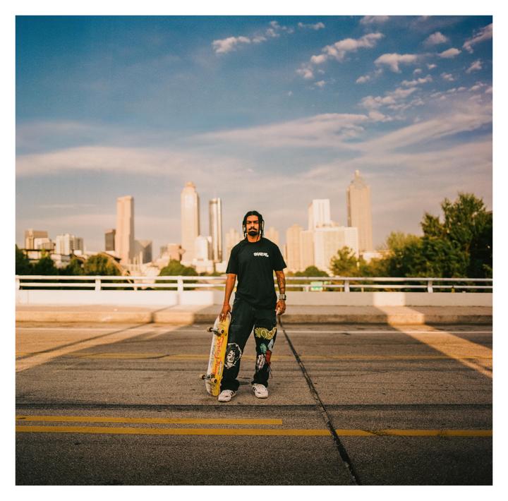 A man stands on a bridge holding a skateboard with a city skyline in the background.