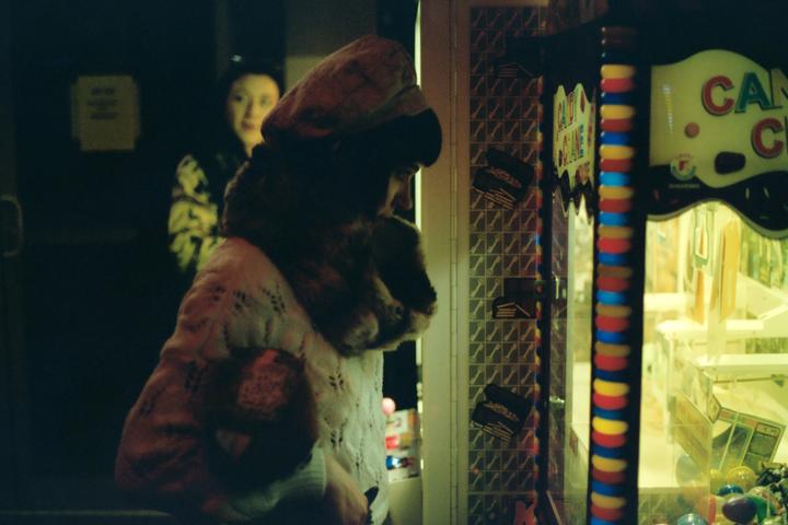 A woman in a hat and fur collar looks into a brightly lit claw machine while another woman stands in the background.