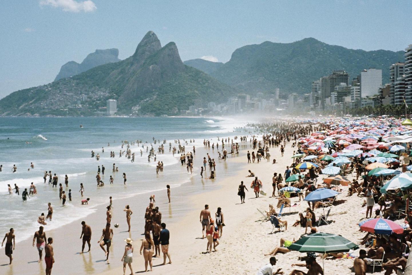 A crowded beach with many people in the water and under colorful umbrellas, with mountains and a city in the background.