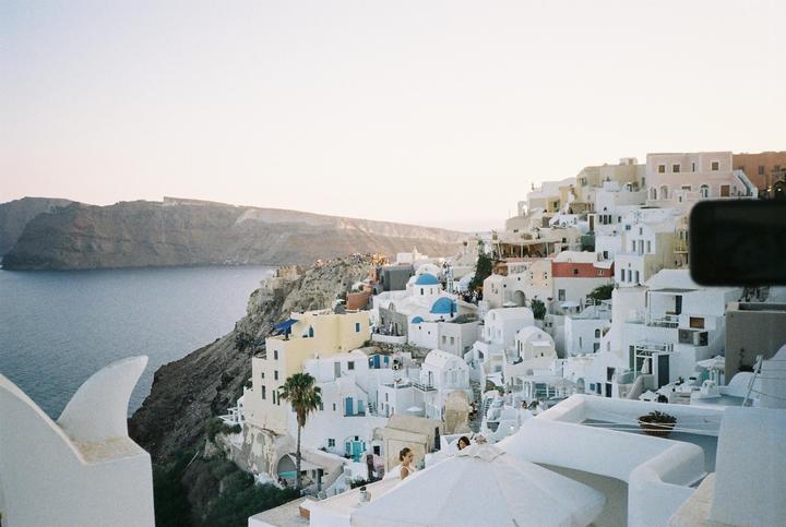 A hillside town with white buildings and blue domes overlooks a body of water and distant mountains.