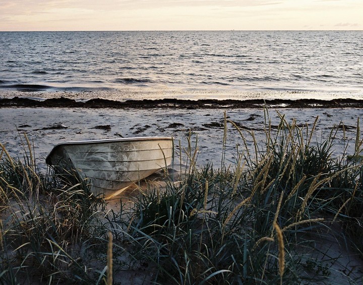 A small boat rests on a sandy beach with tall grass in the foreground and the ocean in the background under a pale sky.